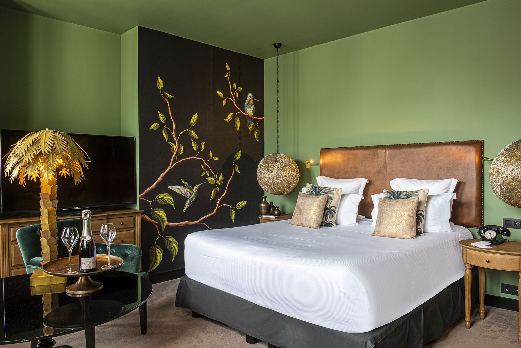 Habitación de hotel con una cama grande, un televisor, una mesita de vidrio con una botella y vasos, y un mural en la pared.