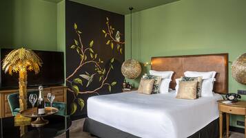 Habitación de hotel con una cama grande, un televisor, una mesita de vidrio con una botella y vasos, y un mural en la pared.