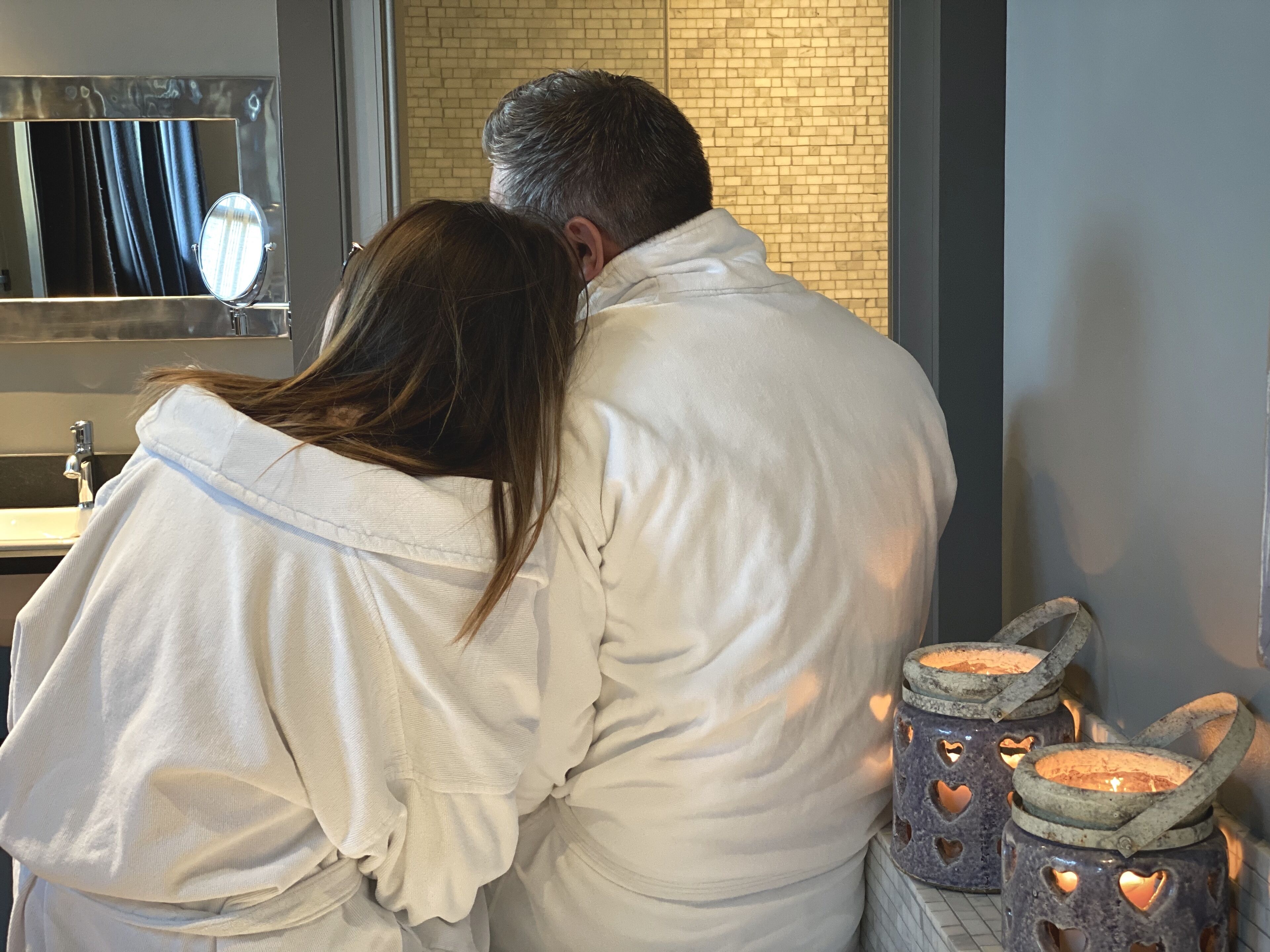A couple embracing in a bathroom with a mirror, sink, and decorative lanterns.