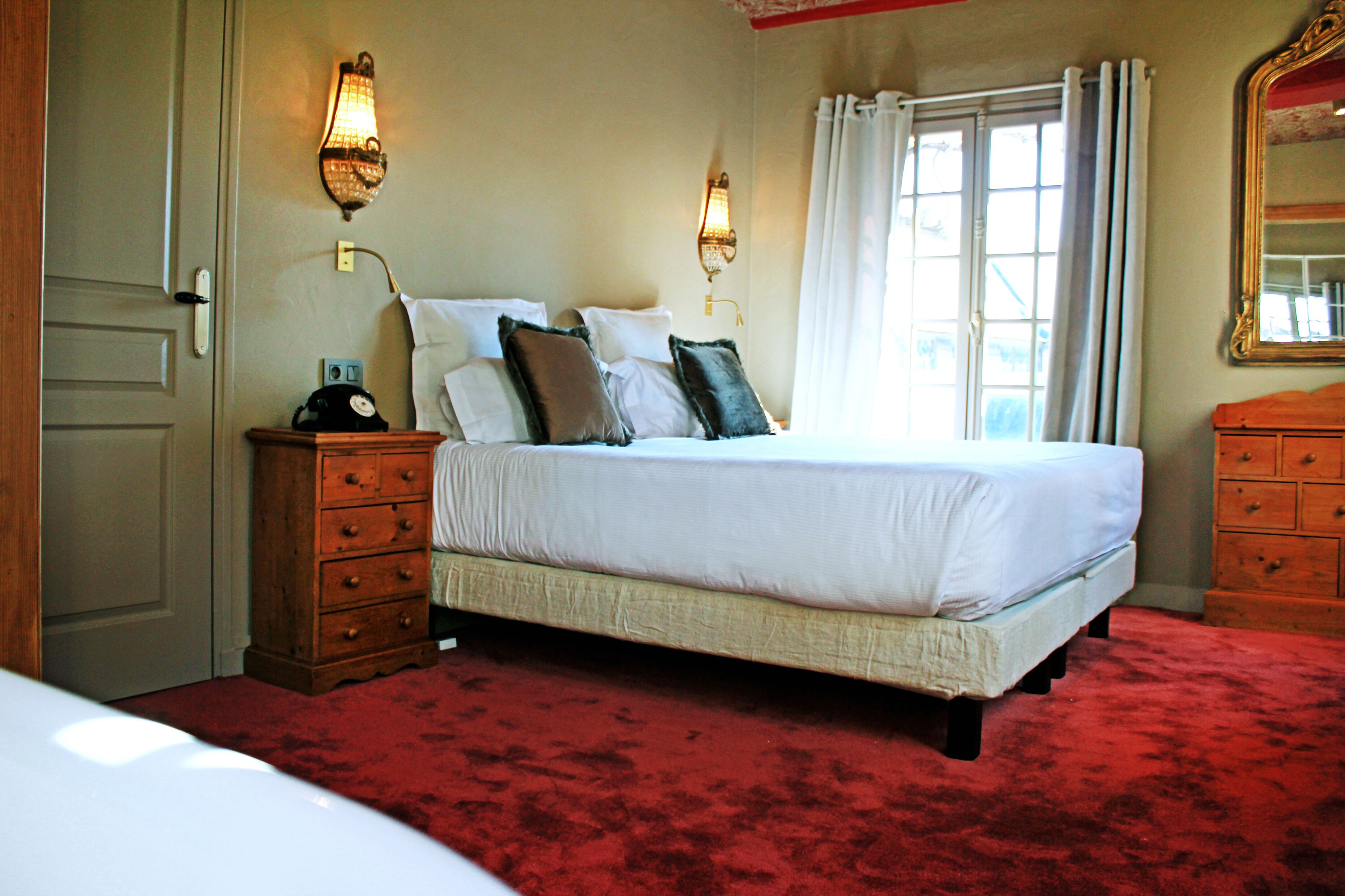 Un dormitorio con una cama grande, mesitas de noche de madera, alfombra roja, dos apliques de pared, una ventana con cortinas y un espejo.