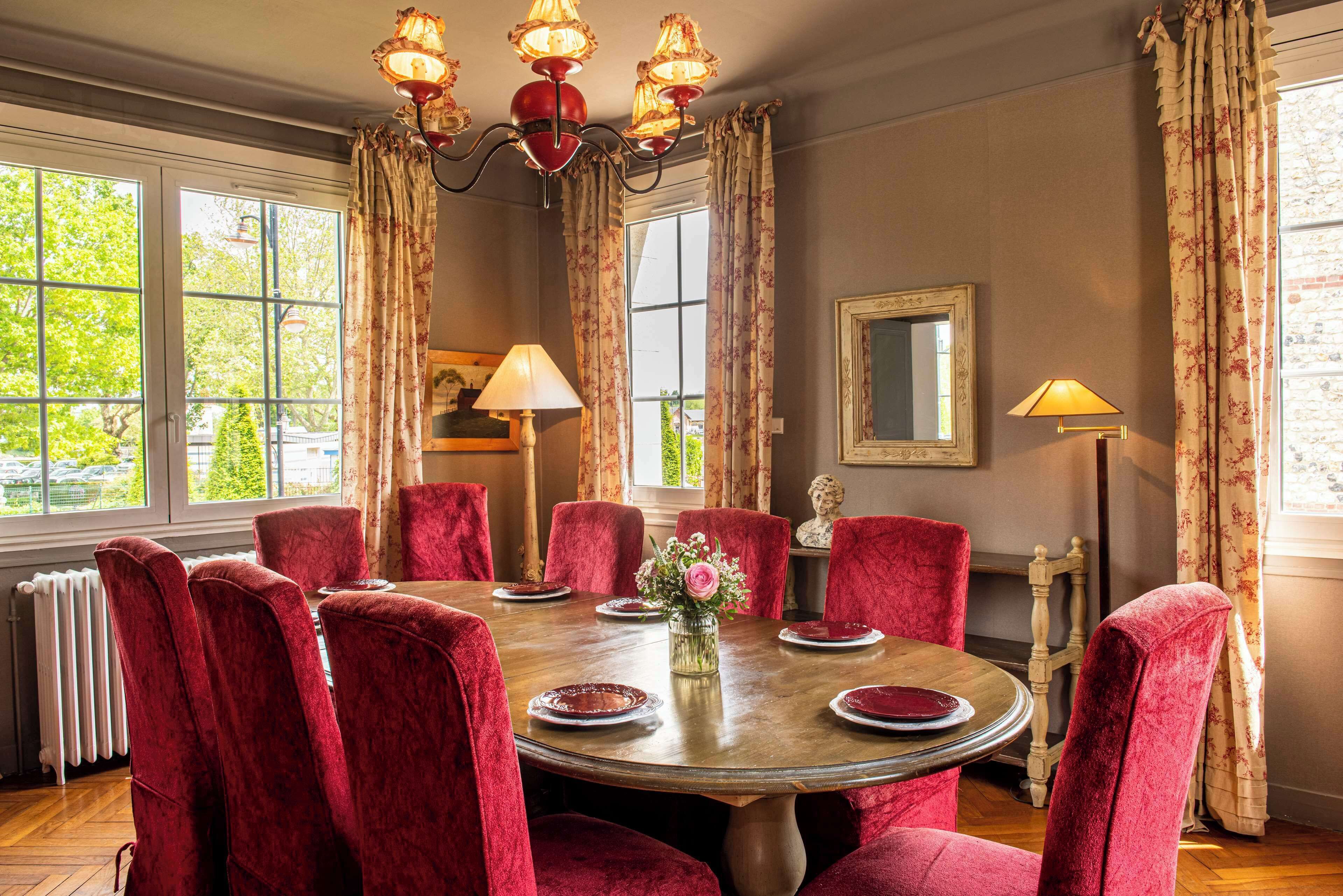 A dining room with a round table, red upholstered chairs, floral curtains, and a chandelier.
