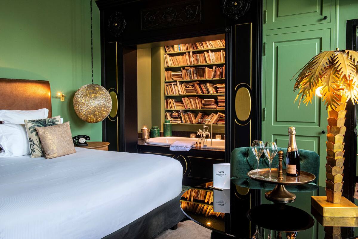 Habitación de hotel con una cama grande, una estantería, una pequeña mesa con vasos y una botella, y una pared verde.