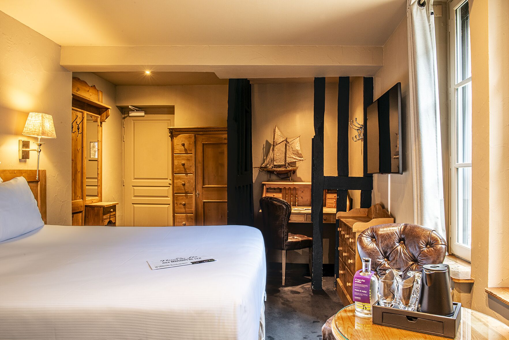 Une chambre d’hôtel avec un grand lit, un bureau orné d’une maquette de bateau, un fauteuil en cuir et une fenêtre avec des rideaux.