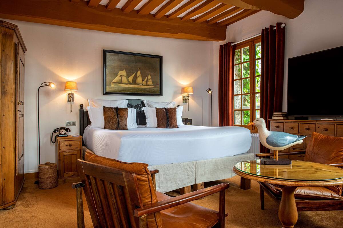 Un dormitorio con una cama grande, dos lámparas de noche, techo de madera, un cuadro sobre la cama, un televisor y una zona de comedor con mesa y sillas.