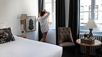 Habitación de hotel con cama, una silla, una mesa con una cesta, una lámpara y una ventana con cortinas.