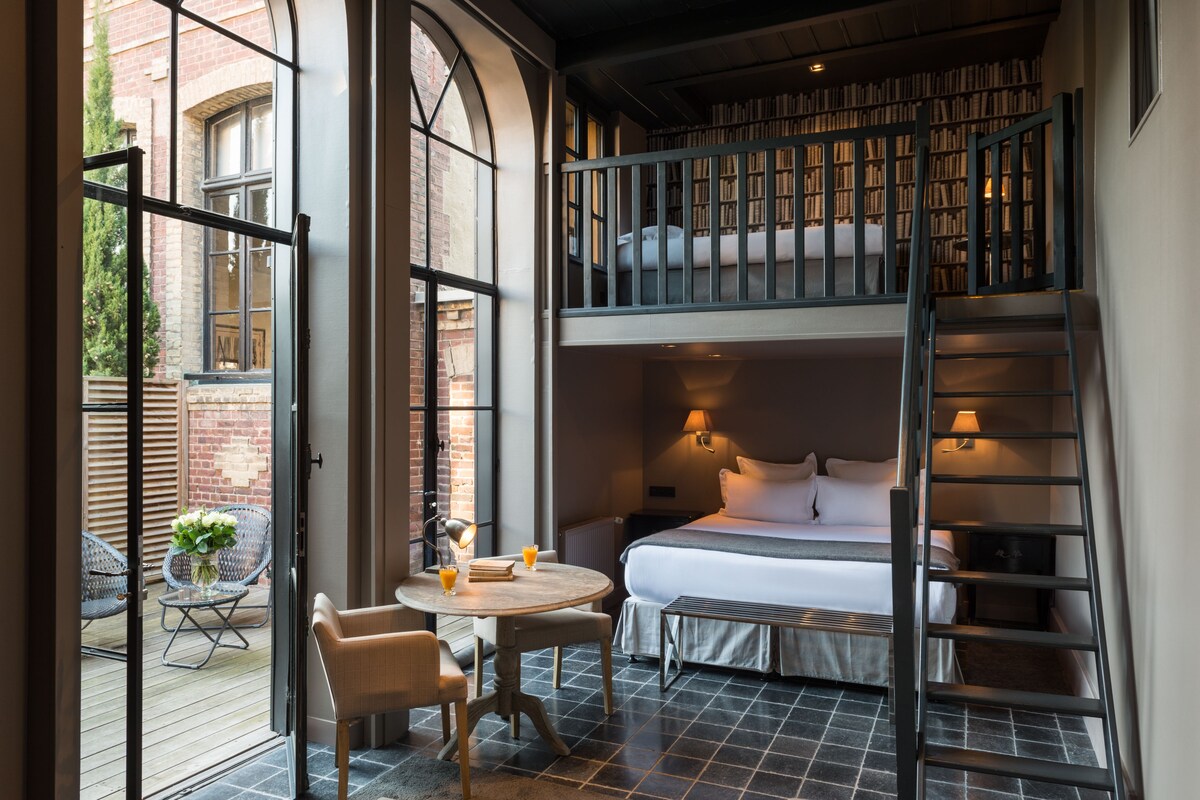 Ein Hotelzimmer mit einem großen Bett, einem Balkon mit Stühlen, einem kleinen Tisch mit Getränken und einem Bücherregal.