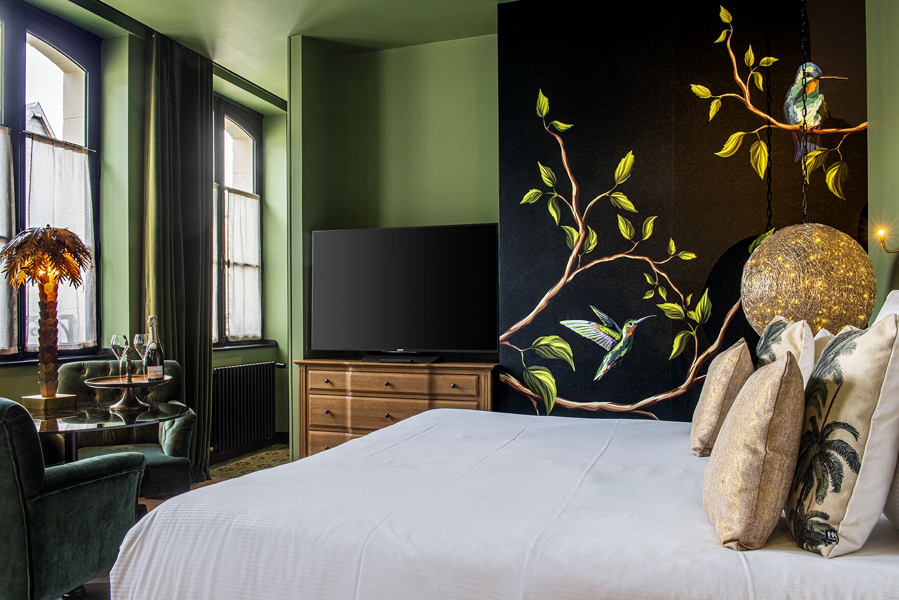 A hotel room with a large bed, a flat-screen TV, a wooden dresser, a dining table with chairs, and a decorative wall with a bird and branch mural.