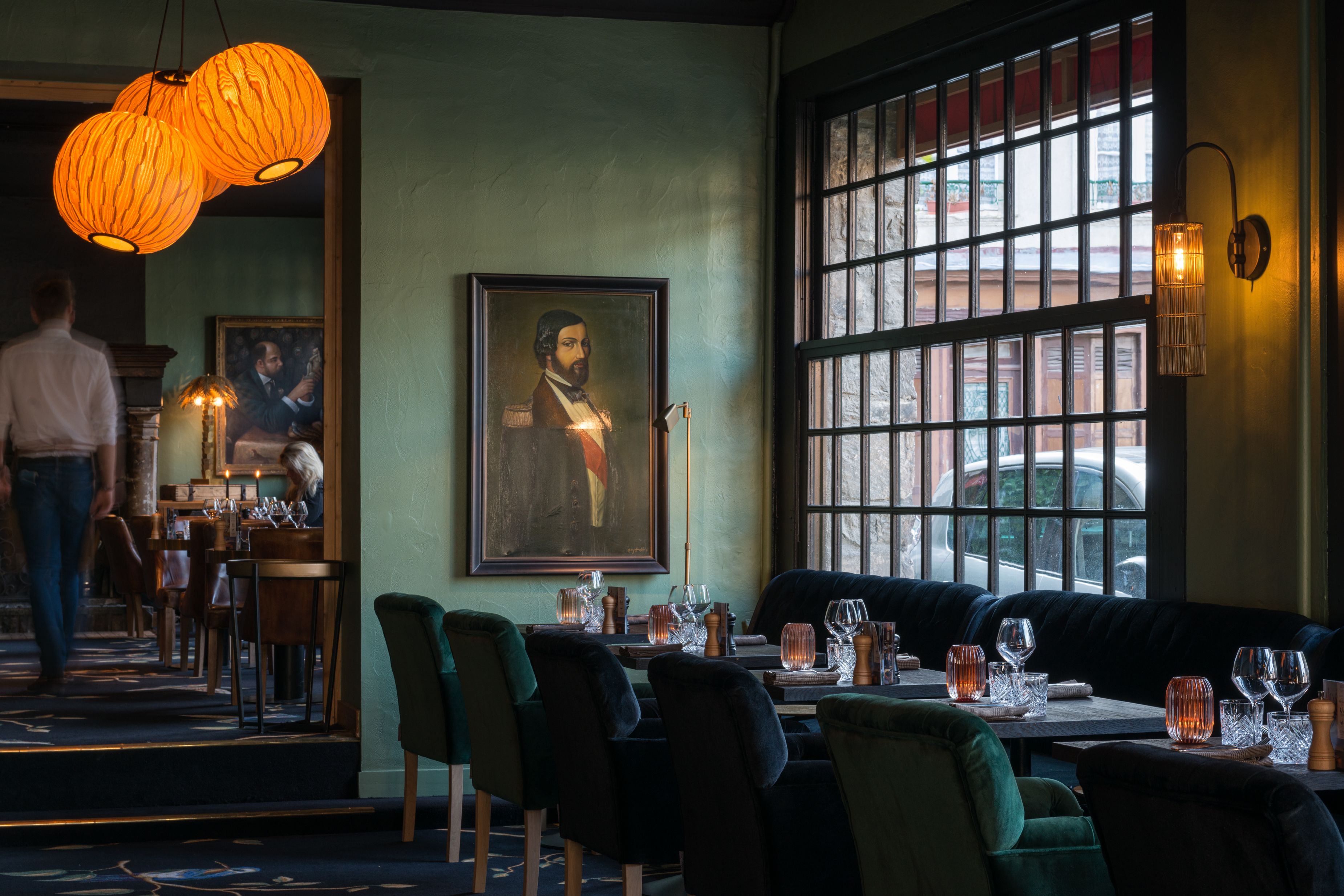 A restaurant interior with a large window, a framed portrait on the wall, and a person walking in the background.