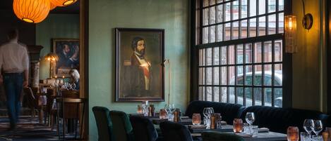 A restaurant interior with a large window, a framed portrait on the wall, and a person walking in the background.