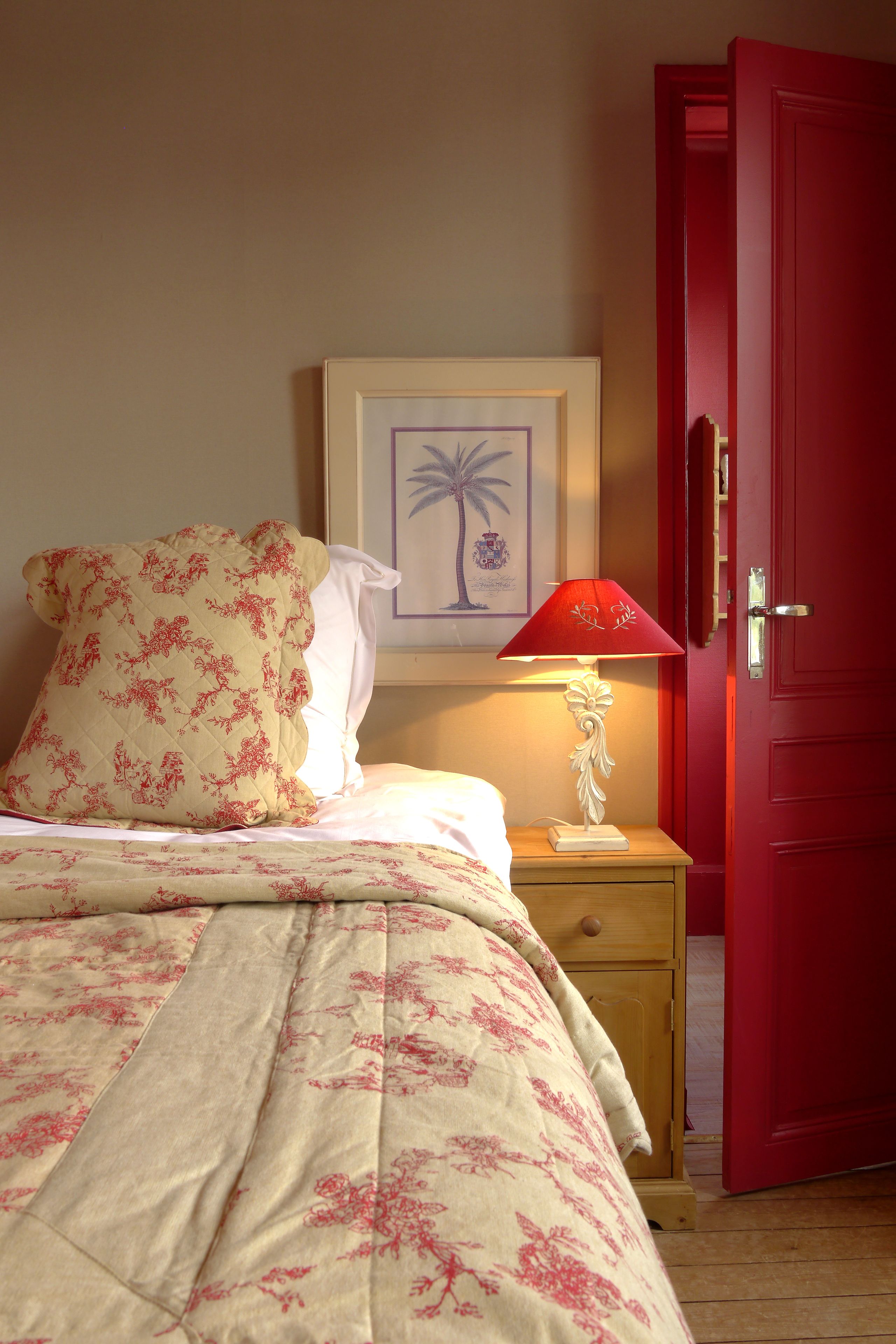 A bed with floral bedding, a white pillow, a red lamp, and a framed picture on the wall.
