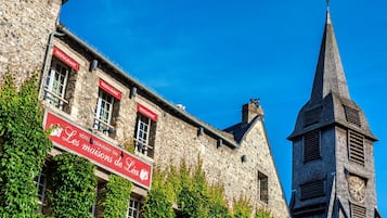 Experience the charm of Les musons de Lia, a historic building with a clock tower and steeply pitched roof, set amidst lush greenery.