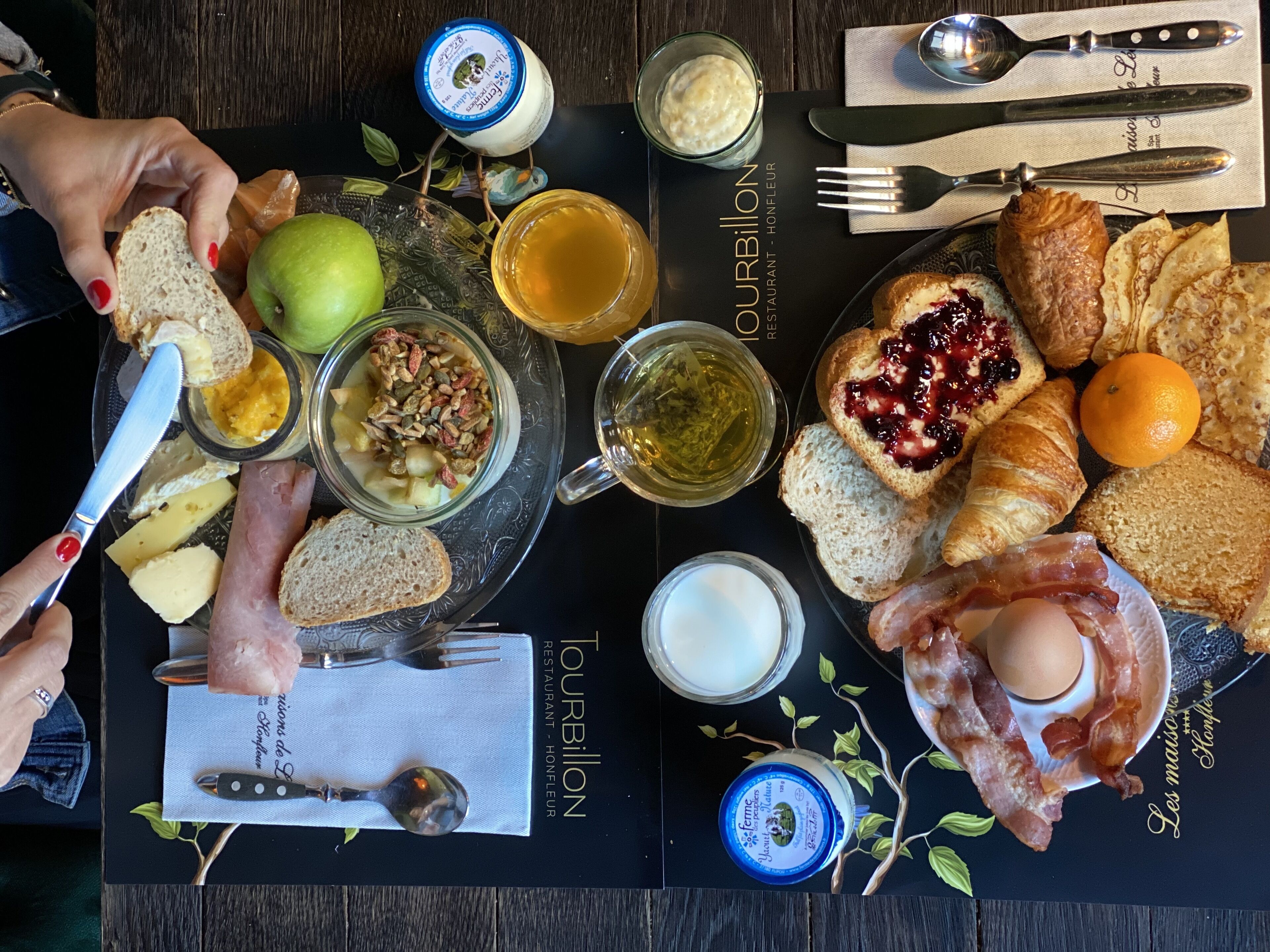 Un assortiment de mets pour le petit-déjeuner est disposé sur une nappe noire, accompagné d'un menu comprenant des toasts, des œufs, du bacon, des viennoiseries et diverses confitures.