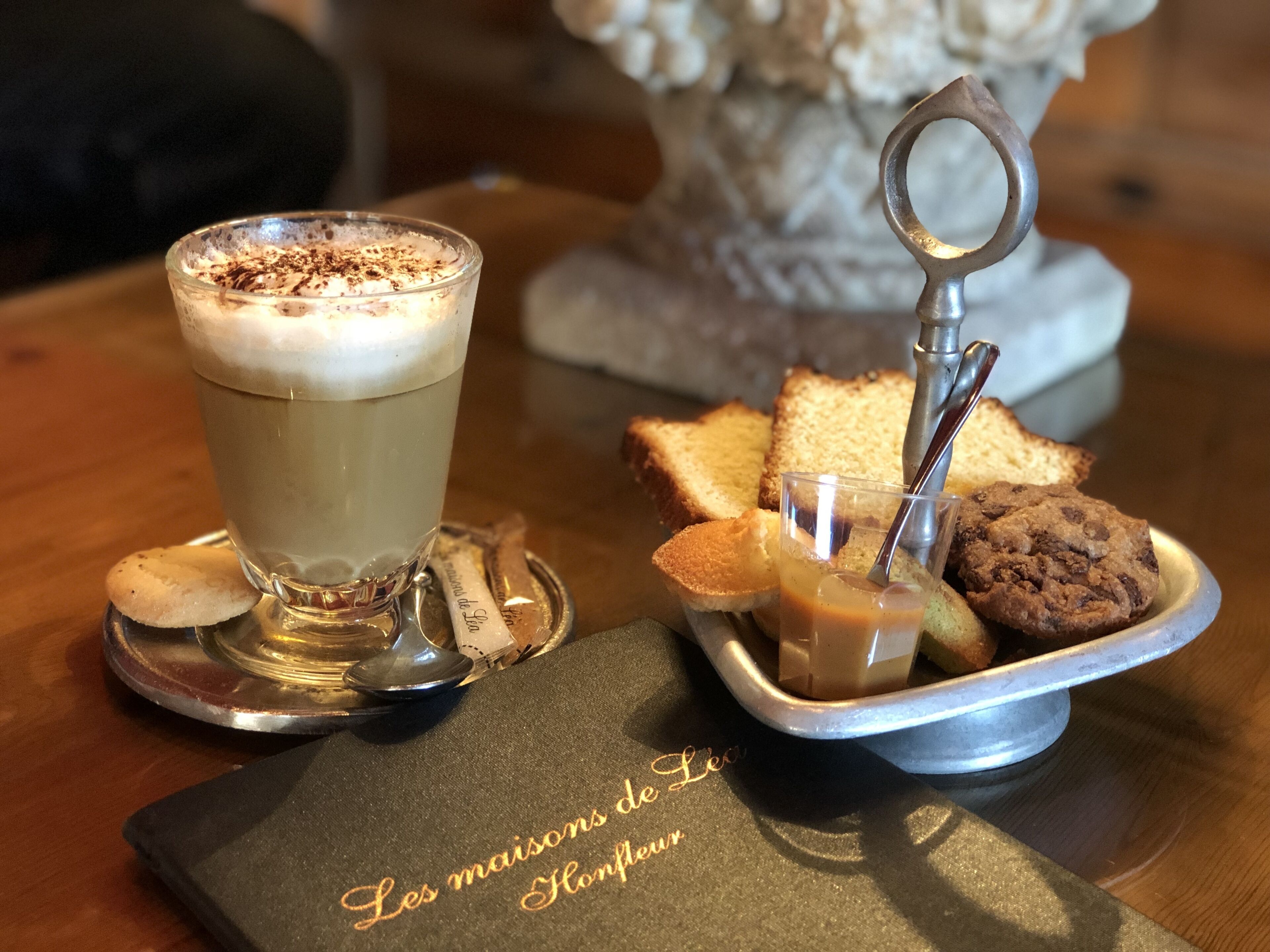 A glass of coffee with foam, a plate of cookies, and a spoon on a coaster.