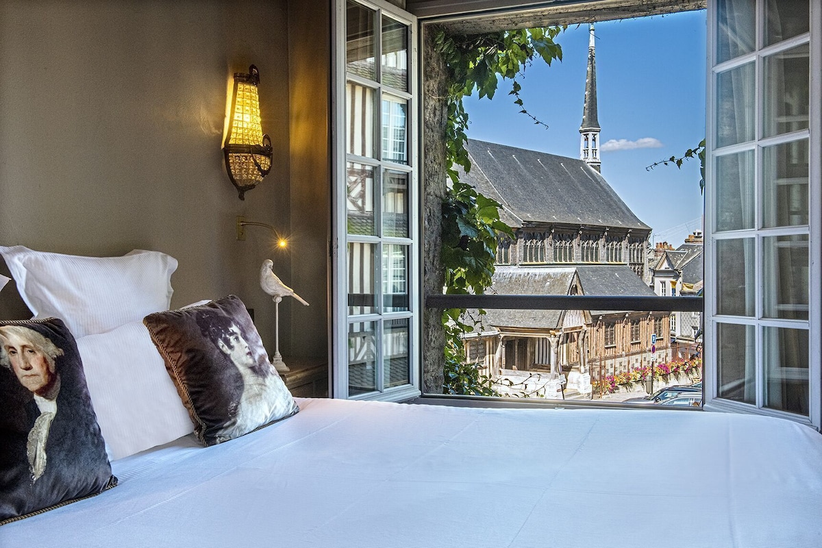 Ein Schlafzimmer mit einem großen Bett, einer Wandlampe und Blick auf ein Gebäude mit einem Turm durch ein Fenster.