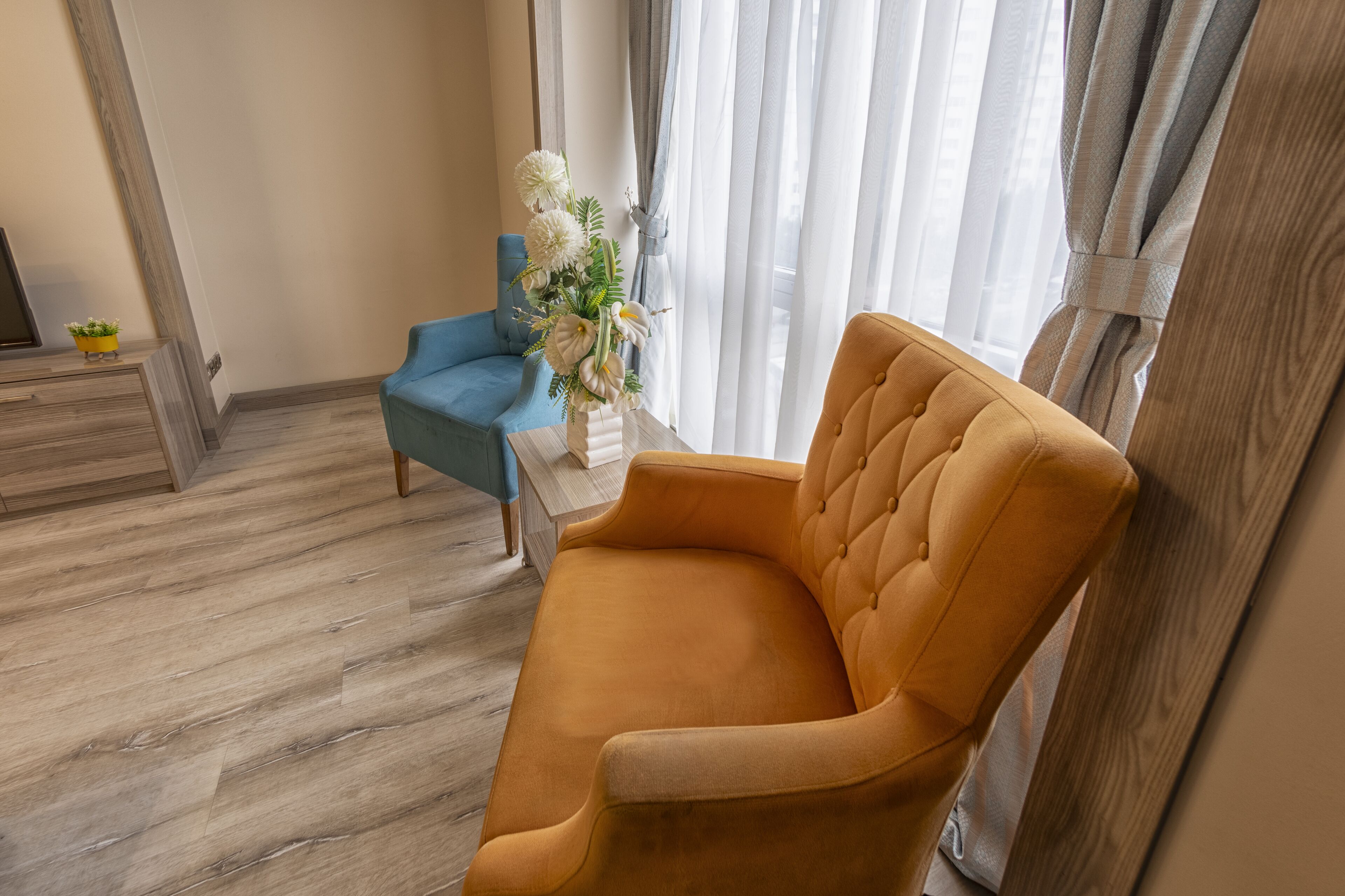Une pièce avec un plancher en bois, un fauteuil bleu, un fauteuil jaune et une petite table sur laquelle est posé un vase de fleurs.