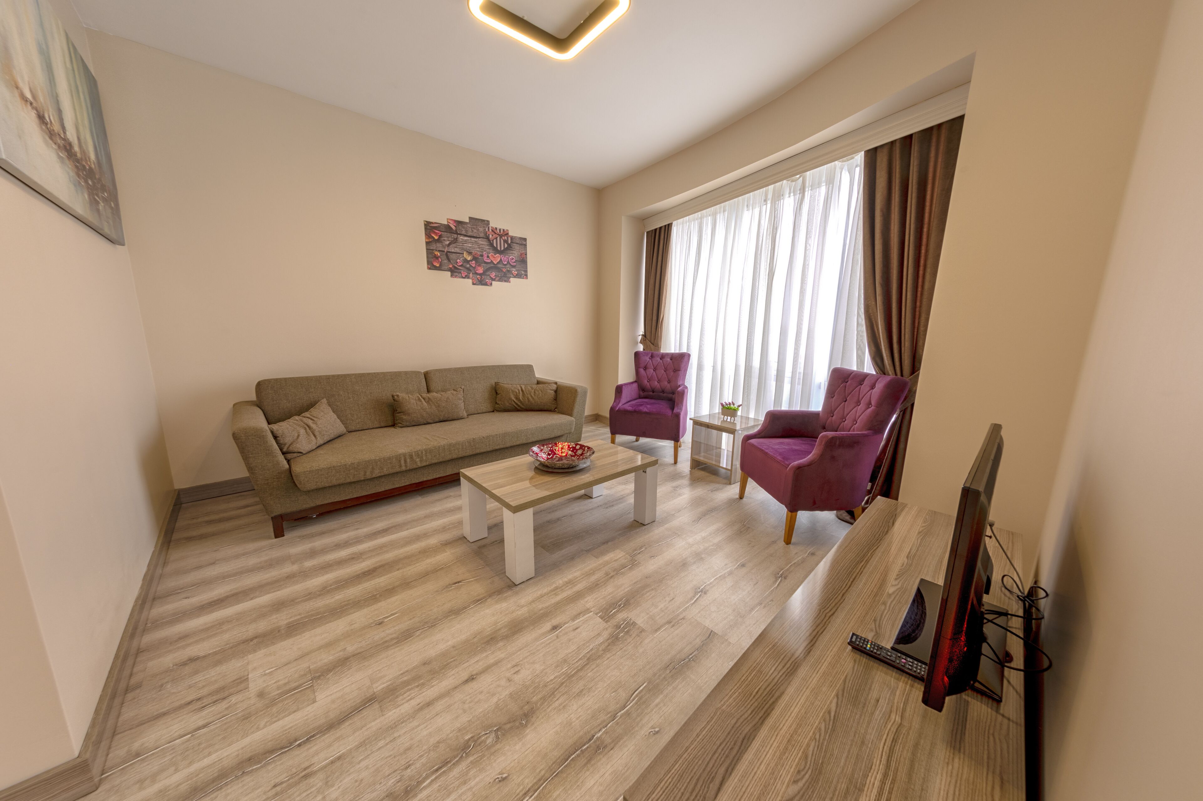 A modern living room with a beige sofa, two purple armchairs, a wooden coffee table, and a flat-screen TV on a wooden stand.