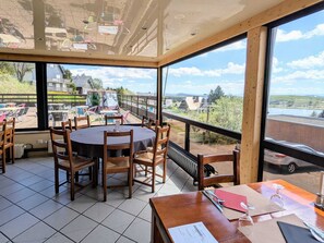 A dining area with a round table and chairs, large windows with a view of a building and landscape, and a wooden ceiling. - Hôtel Gergovia (Besse-et-Saint-Anastaise)