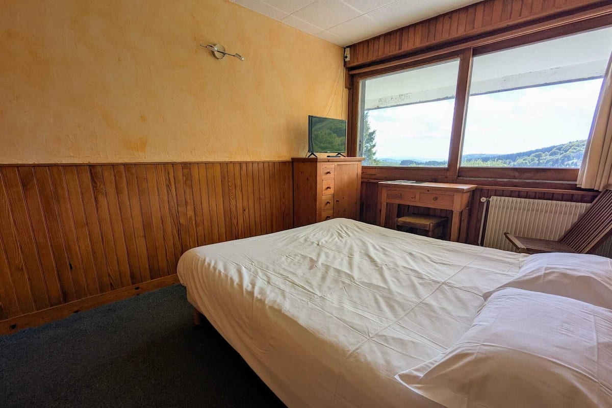 A bedroom with a bed, a wooden desk, a chair, and a window with a view.