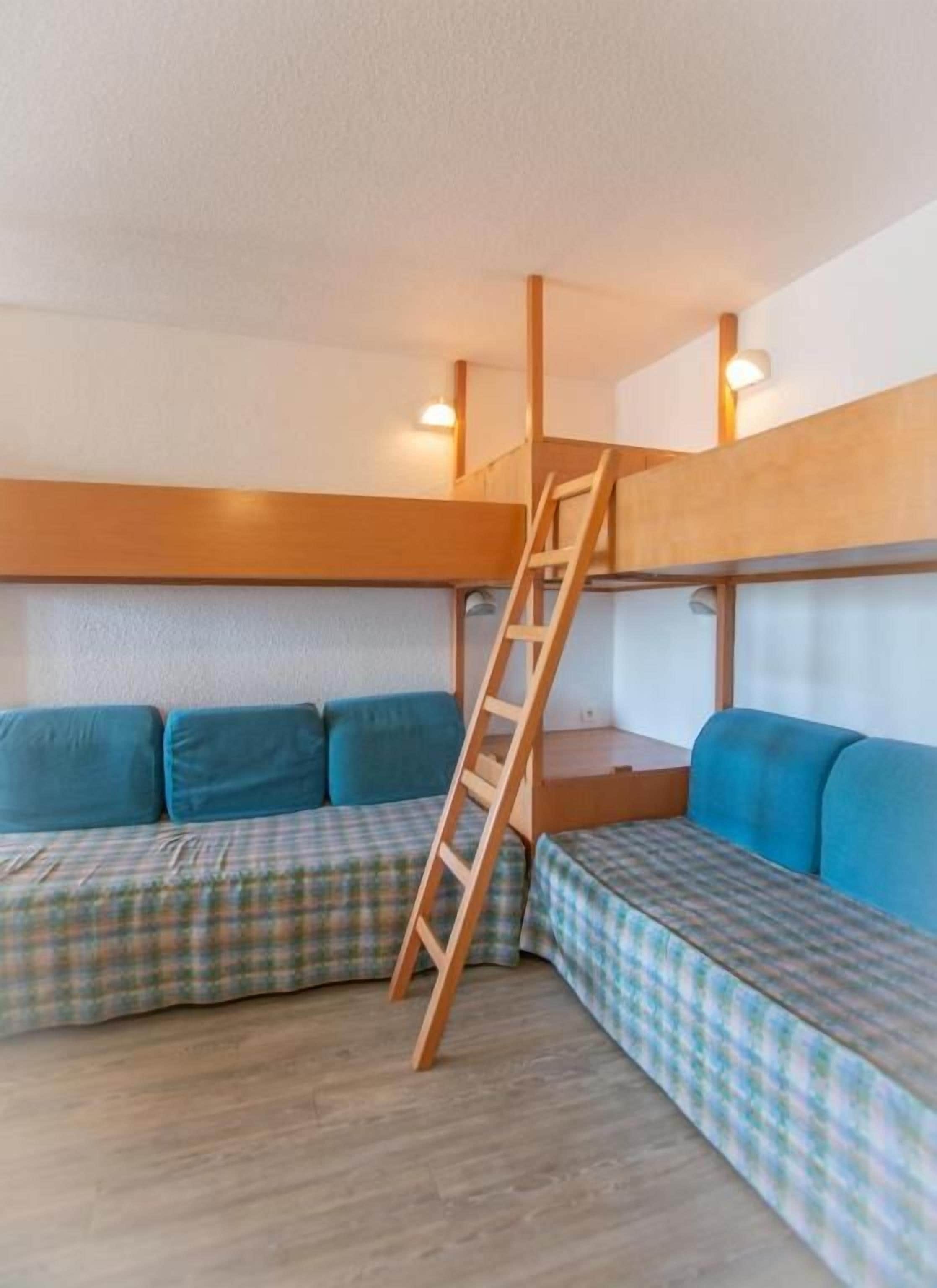 Une chambre avec un lit superposé et une échelle, deux bancs bleus recouverts de tissu et un plancher en bois.