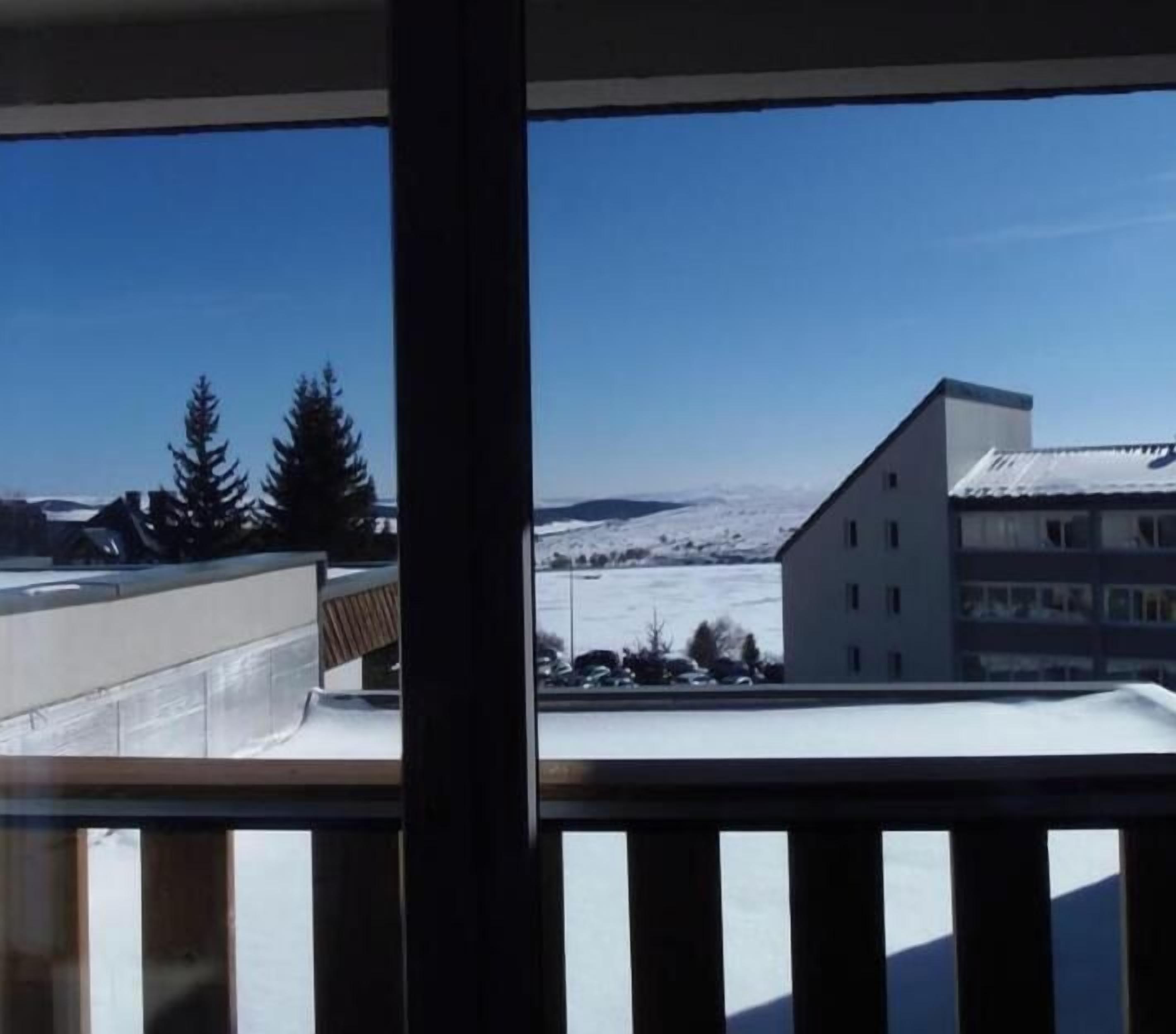 A view from a window with a balcony, overlooking a building with a sloped roof and a snow-covered landscape.
