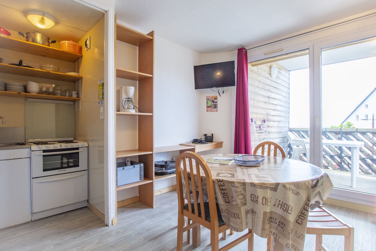 Une cuisine compacte équipée d’une cuisinière, d’un four et d’étagères où sont rangés la vaisselle. On y trouve également une table à manger avec des chaises et un téléviseur fixé au mur.
