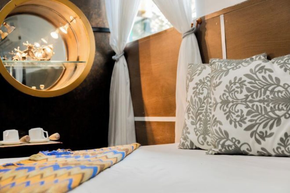 A hotel room with a bed, a round mirror, a bedside table with cups and a decorative item, and patterned pillows.
