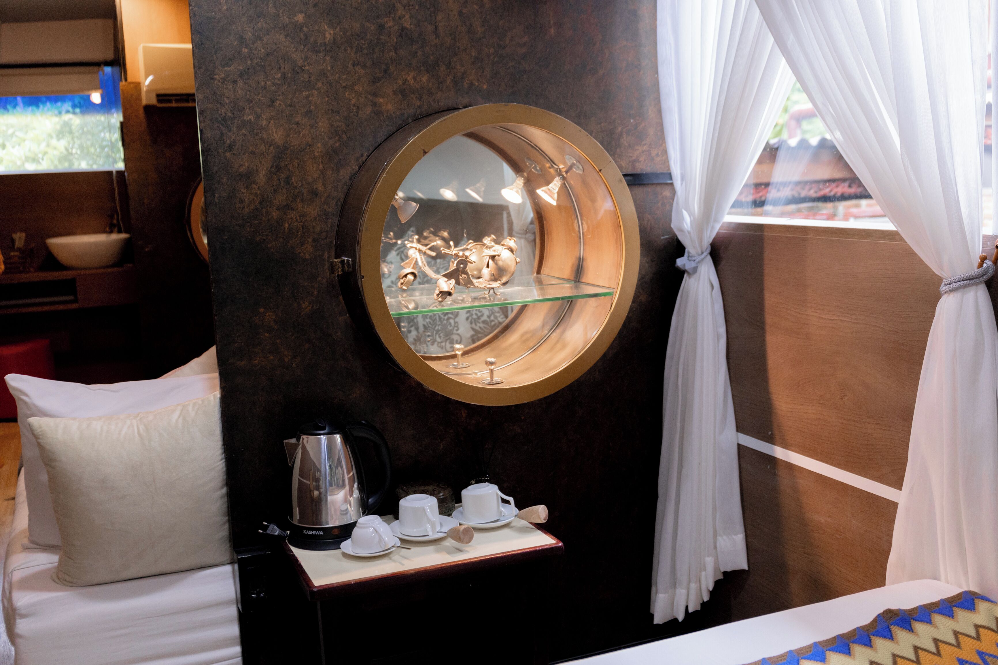 Une chambre d’hôtel avec un miroir rond, un lit avec une literie blanche, une petite table avec un service à thé, une machine à café et une vue sur l’extérieur à travers des rideaux transparents.