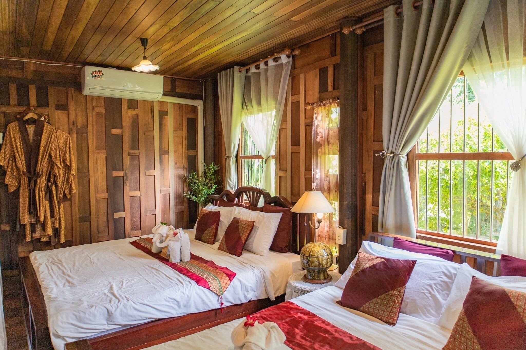 A room with two beds, wooden walls, a hanging towel rack, a globe lamp, and a window with curtains.