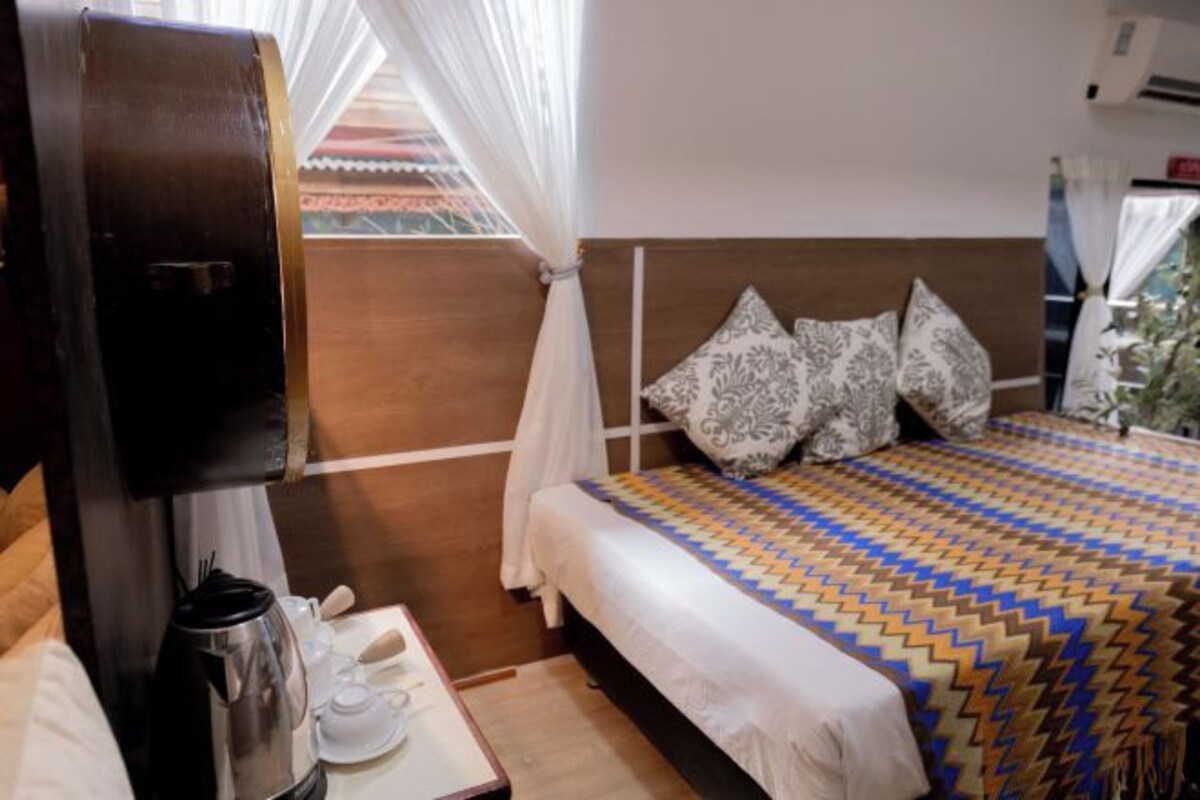 A hotel room with a bed, a bedside table with a kettle, cups, and a tray, a wooden headboard, patterned pillows, and a view of the outside through a window with sheer curtains.