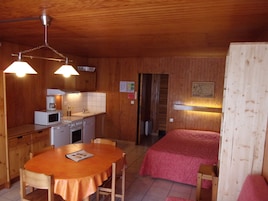 Habitación compacta con paredes de madera y una kitchenette. Cuenta con una mesa de comedor con sillas, una cama con una manta rosa y un baño pequeño.