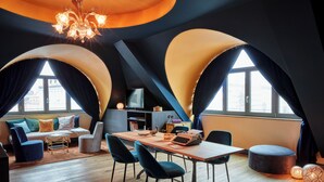 Een moderne woonkamer met een bank, stoelen, een eettafel en een televisie. Er zijn grote ramen met gordijnen, een kroonluchter en een open haard.