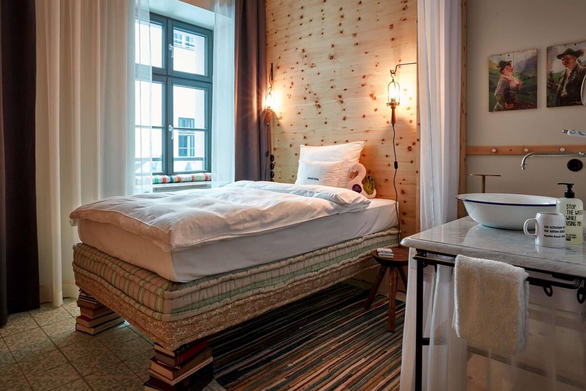 A hotel room with a bed, a bedside table with a bottle, a window with curtains, a wooden wall, and a bathroom with a sink and towel rack.