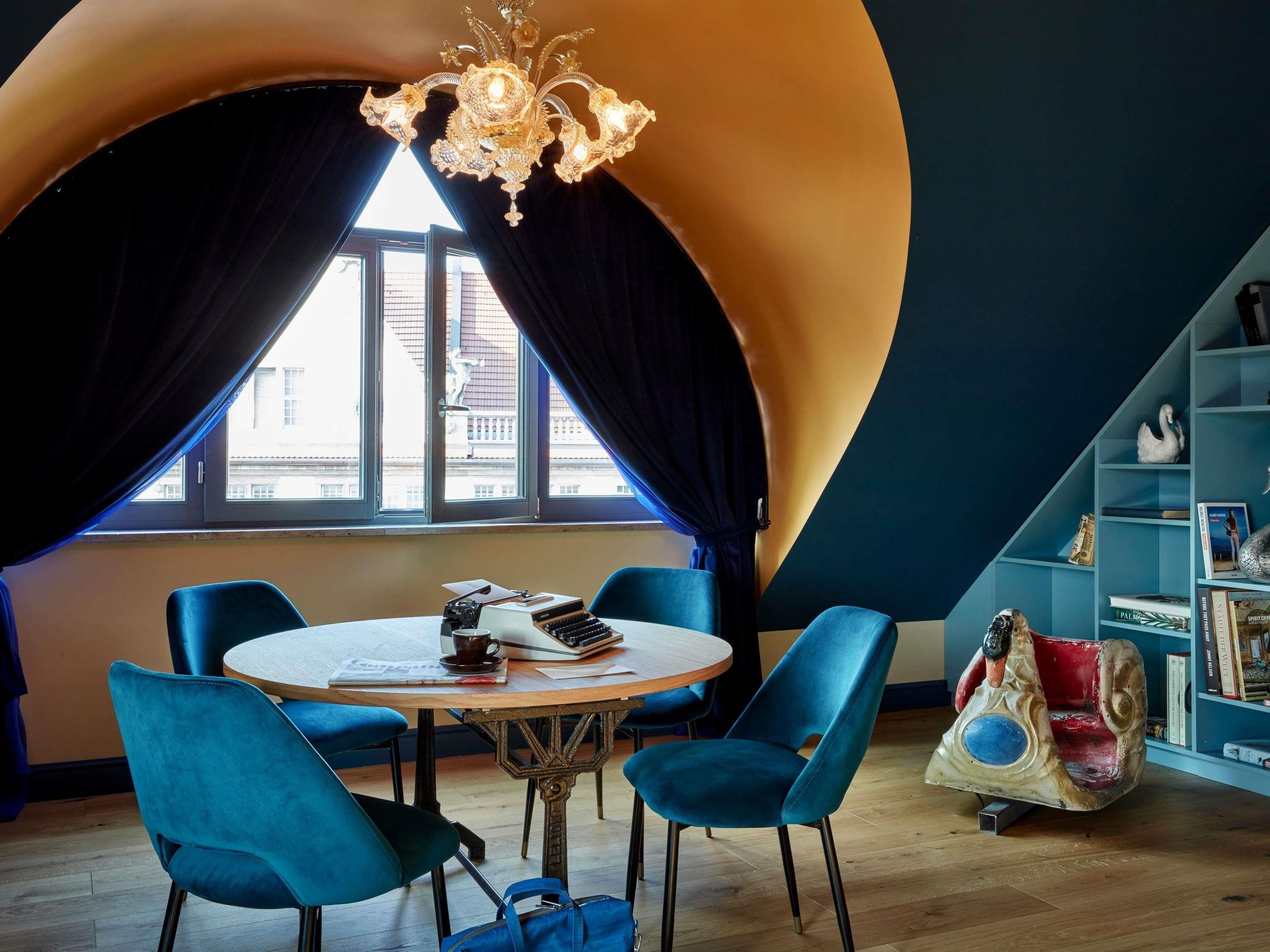 A room with a round table, blue chairs, a chandelier, a bookshelf, and a view through a window with curtains.