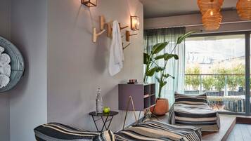 A modern living room with a sofa set, a side table with a bottle and apple, wall-mounted lights, and a large window with a view.