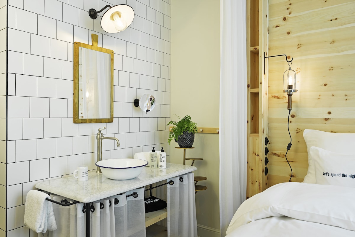 Ein modernes Badezimmer mit weiß gefliester Wand, einem мраморным Waschbecken, einem Spiegel, einem Handtuchhalter, einer kleinen Ablage mit Pflegeprodukten und einer Topfpflanze.