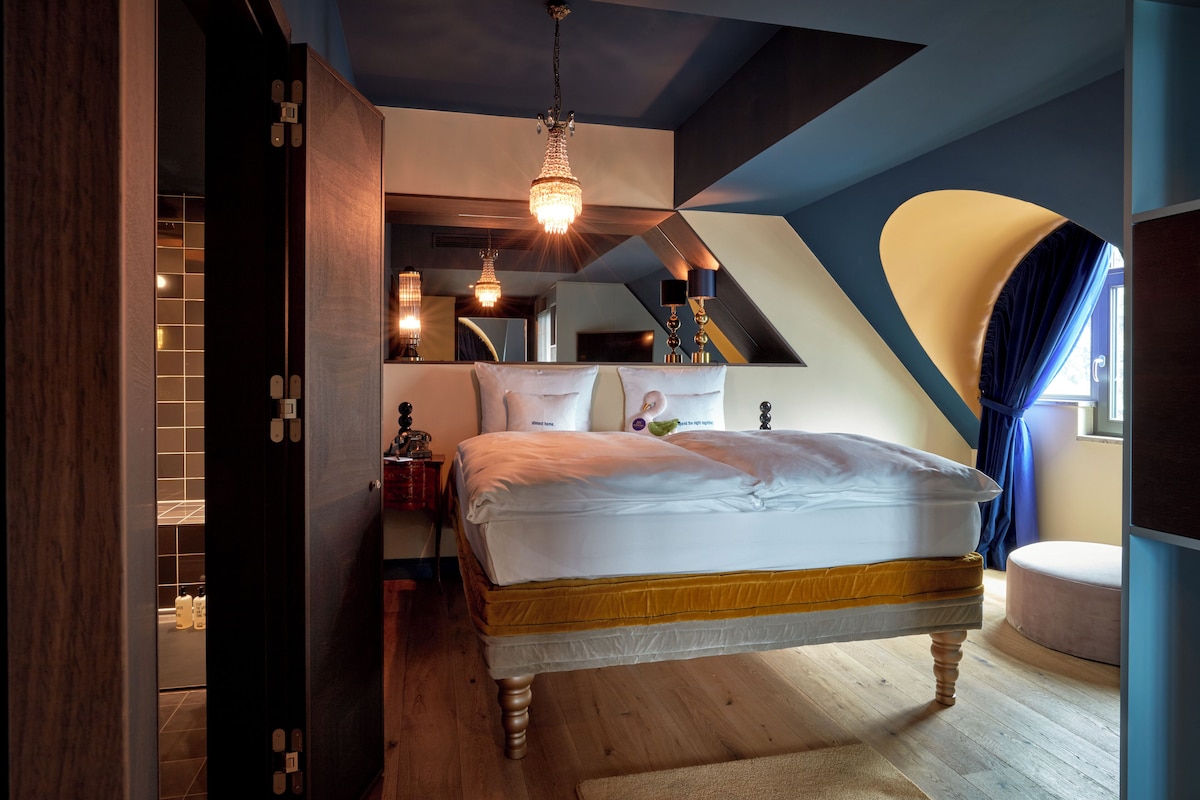 Ein Schlafzimmer mit einem großen Bett, Holzboden, einer Kristallleuchtre und einem Fenster mit Vorhängen.