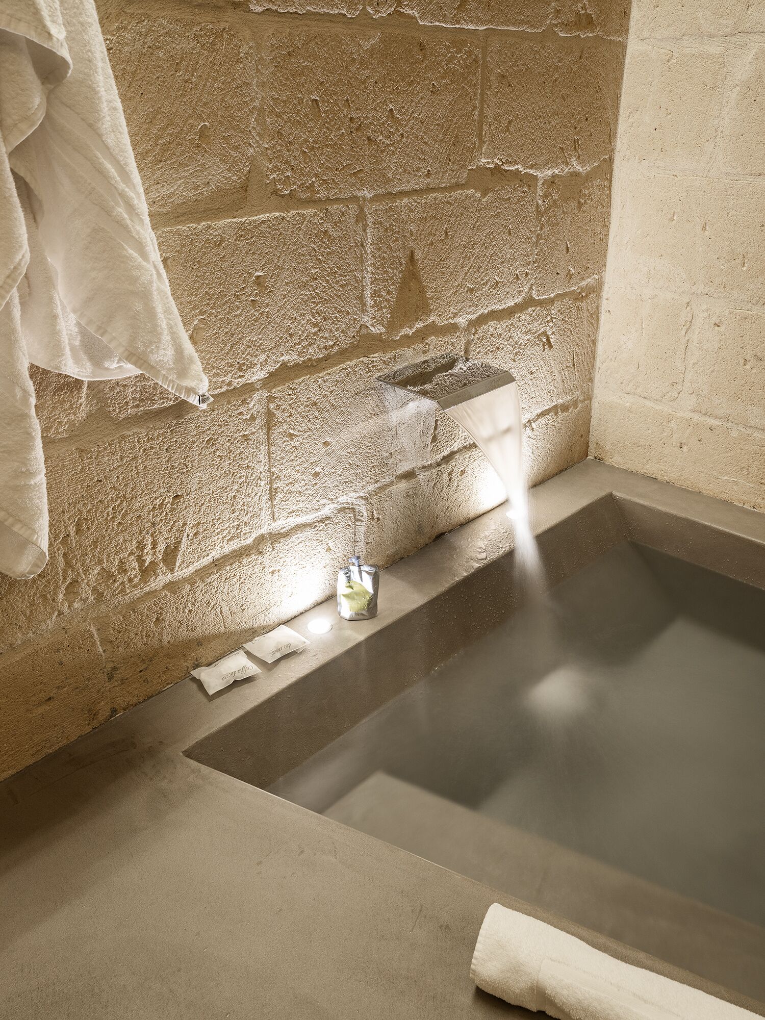 Ein Badezimmer mit Steinwänden, einer Badewanne, einem an der Wand hängenden Handtuch und einer kleinen Flasche auf dem Boden.