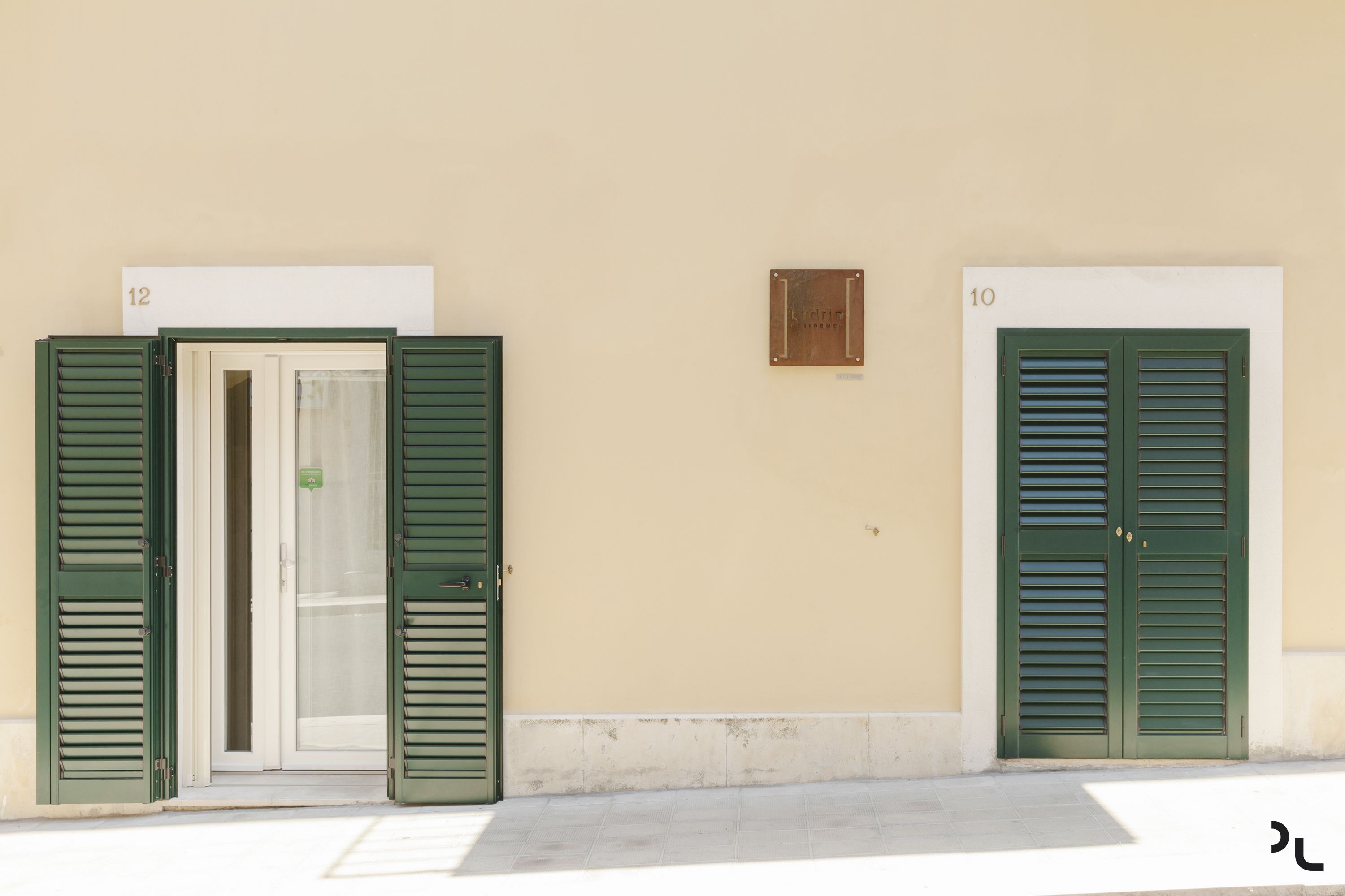 Zwei Türen mit grünen Fensterläden, mit den Hausnummern 10 und 12, an einer beigen Wand.