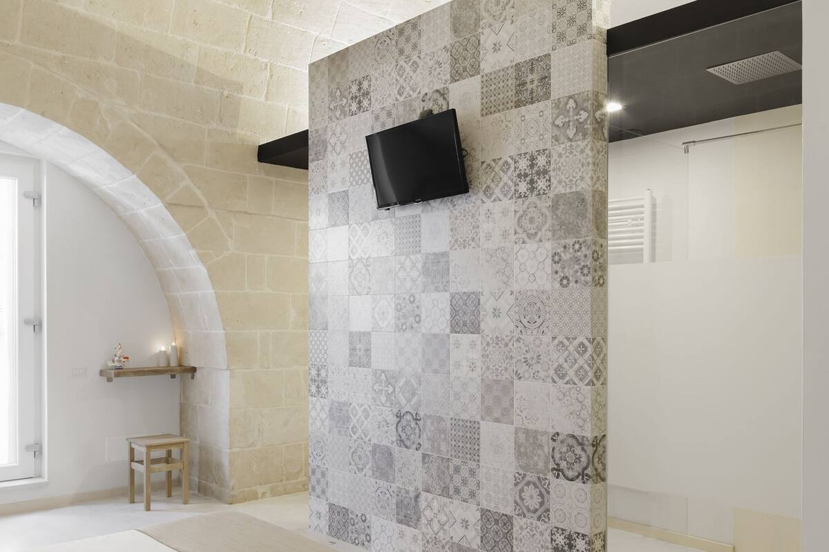 A modern hotel room with a large bed, a TV mounted on a patterned wall, and a small seating area with a chair and shelf.