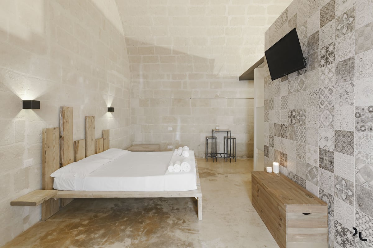 A bedroom with a wooden bed, a wooden bench, a TV mounted on the wall, and a tiled wall with a pattern.