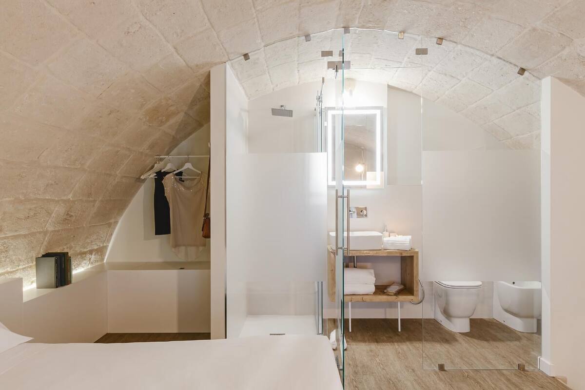 A modern, compact bedroom with a sloped ceiling, a glass-enclosed walk-in closet, and a minimalist bathroom with a wooden vanity.