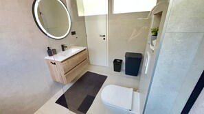 A modern bathroom with a white sink, mirror, and toilet. There is a black trash bin and a small potted plant on the right. - Guesthouse Civljak (Baska Voda)