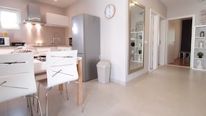 Eine moderne Küche mit weißen Schränken, einer Mikrowelle und einem Kühlschrank. Ein Essbereich mit weißen Stühlen und einem Tisch ist vorhanden. Auf der Wand hängt eine Uhr, und ein Flur führt zu einem Raum mit Regalen.