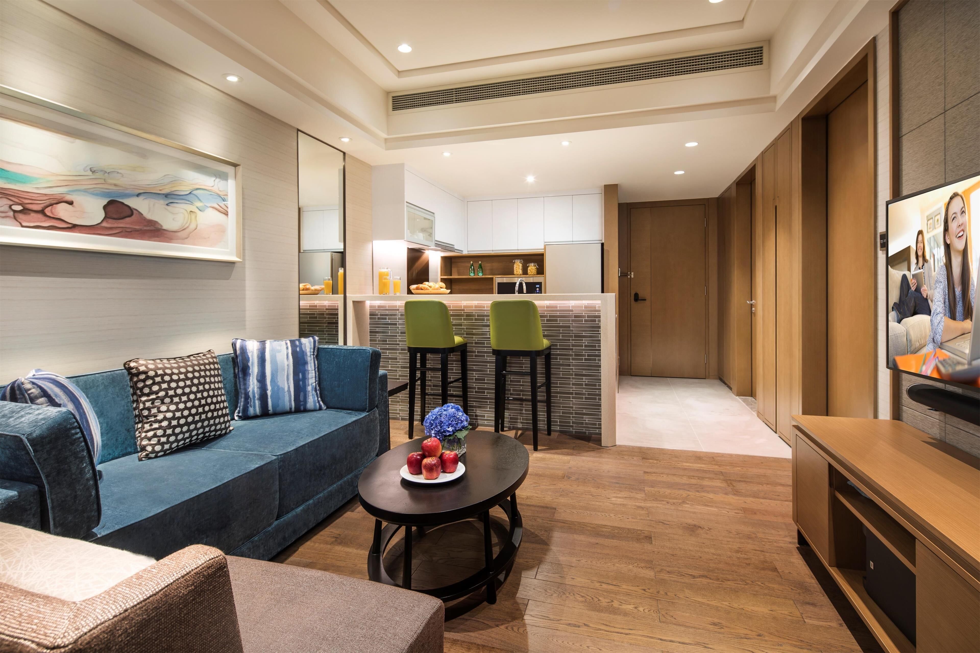 Una sala de estar moderna con un sofá, una mesa de centro y un televisor. Se observa una cocina al fondo con una barra.