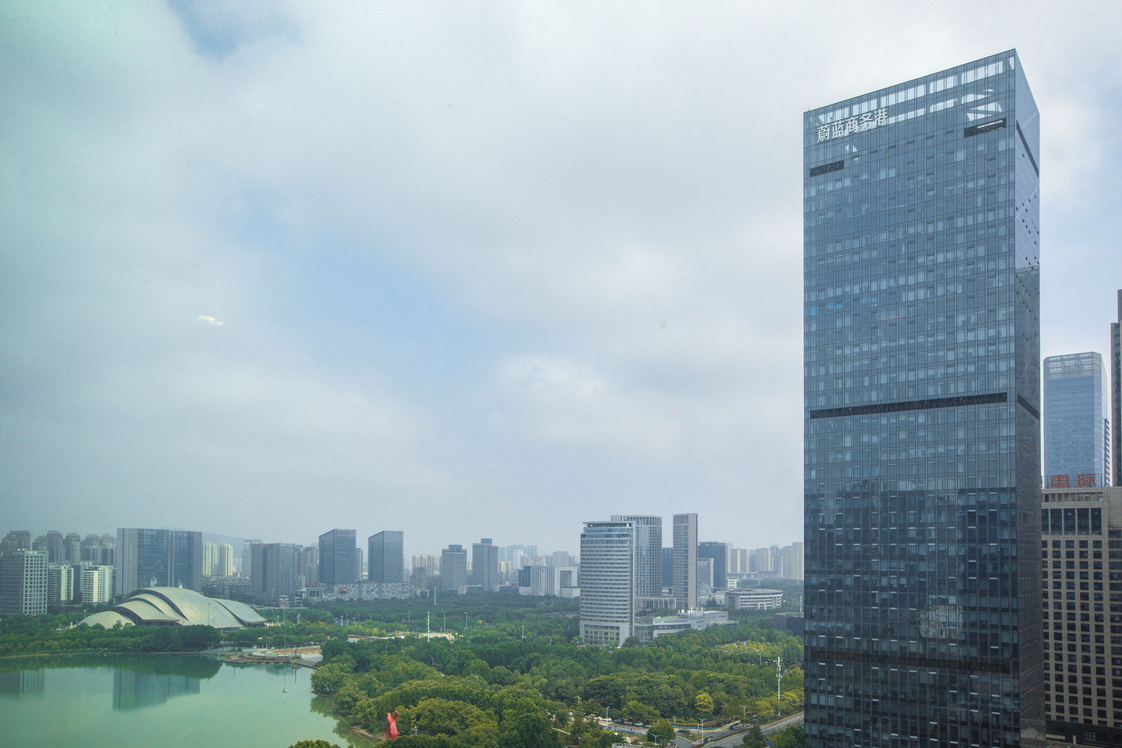 Un immeuble de grande hauteur à la façade vitrée, entouré d’autres gratte-ciel et bordé par un plan d’eau, avec une végétation luxuriante au premier plan.