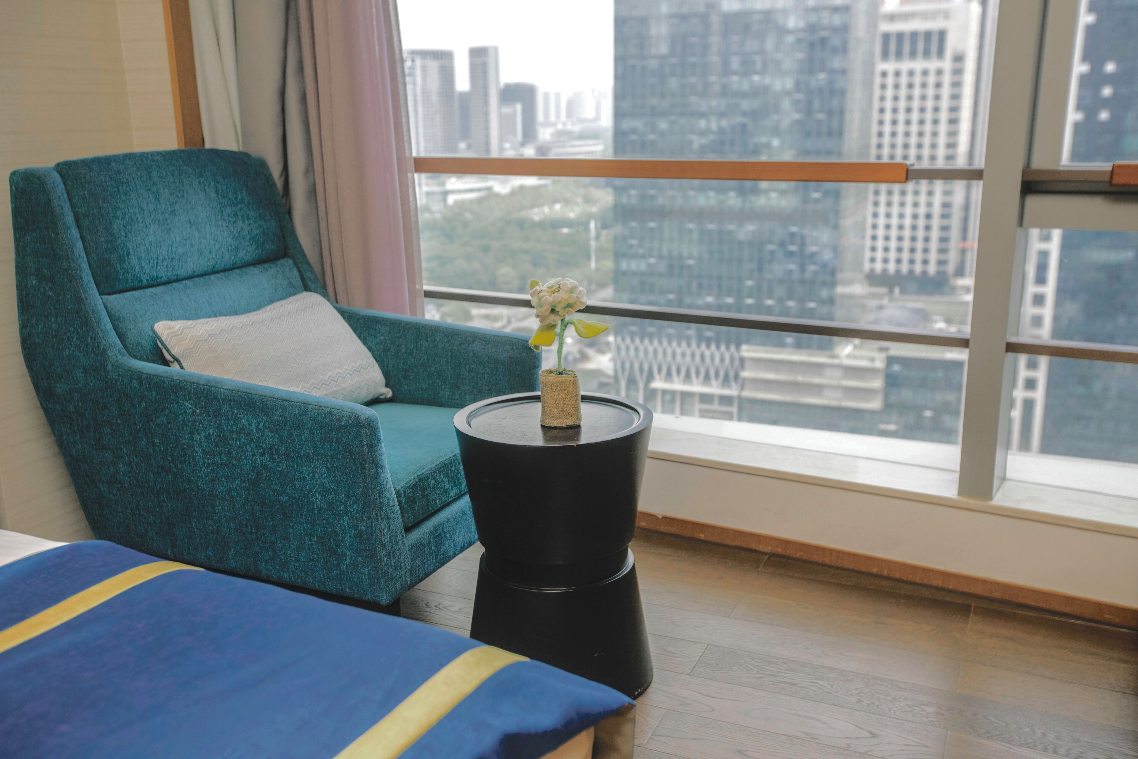 A teal armchair with a white pillow, a small black side table with a vase of flowers, and a view of a cityscape through large windows.