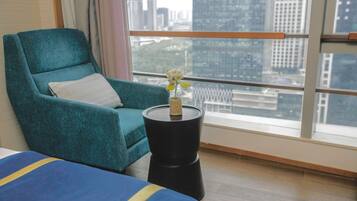 A teal armchair with a white pillow, a small black side table with a vase of flowers, and a view of a cityscape through large windows.