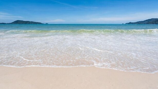 Ein Strand mit klarem Wasser, einem Sandstrand und in der Ferne Hügel.