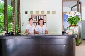 Un comptoir d’accueil avec deux employés, une plante et une bibliothèque en arrière-plan.