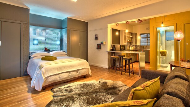 A modern bedroom with a bed, a sofa, and a dining area with a kitchen in the background. - Langholm (Cape Town)
